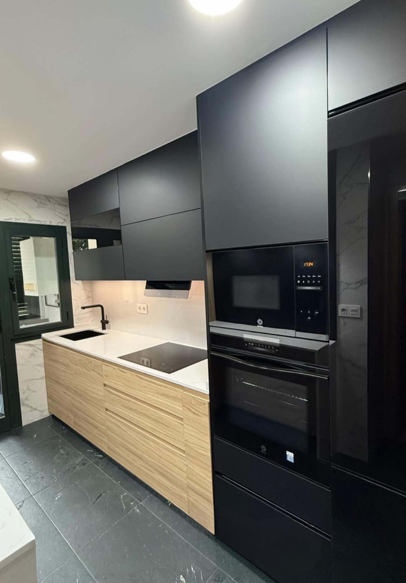 Cozinha renovada em Almada: armários pretos mate, frentes de gaveta em madeira, bancada clara com iluminação de trabalho e eletrodomésticos encastrados pretos