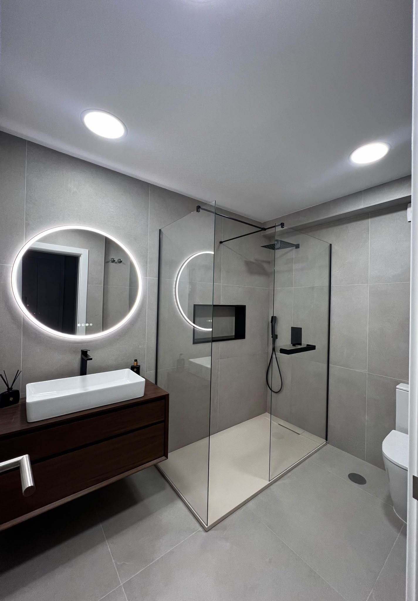 Casa de banho renovada: duche walk-in com resguardo de vidro e perfis pretos, espelho redondo retroiluminado e móvel suspenso em madeira